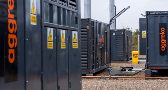 A close-up view of a row of dark gray, containerized Aggreko industrial units, featuring yellow hazard symbols on the sides. The units are installed outdoors on a gravel lot.