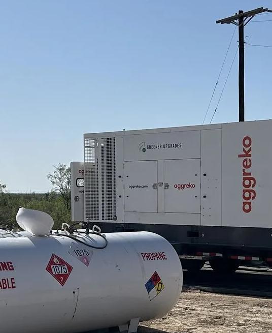 A row of large, white horizontal propane or fuel storage tanks in the foreground. Behind them, several white, containerized Aggreko units are parked on trailers, set against a bright, dry landscape.