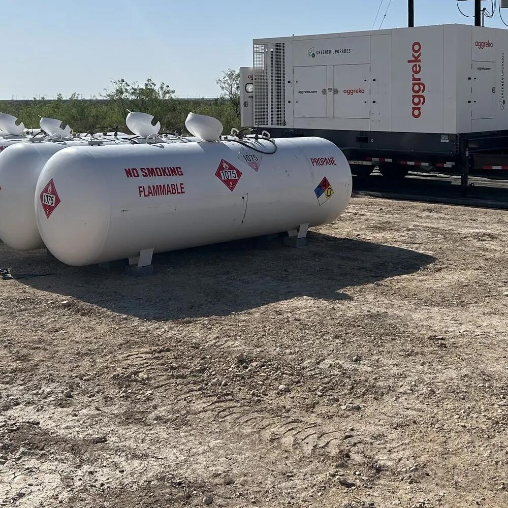 A row of large, white horizontal propane or fuel storage tanks in the foreground. Behind them, several white, containerized Aggreko units are parked on trailers, set against a bright, dry landscape.