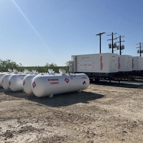 A row of large, white horizontal propane or fuel storage tanks in the foreground. Behind them, several white, containerized Aggreko units are parked on trailers, set against a bright, dry landscape.