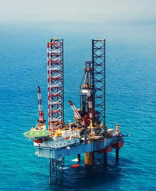 Offshore oil drilling platform with cranes, equipment, and support vessels in blue water.