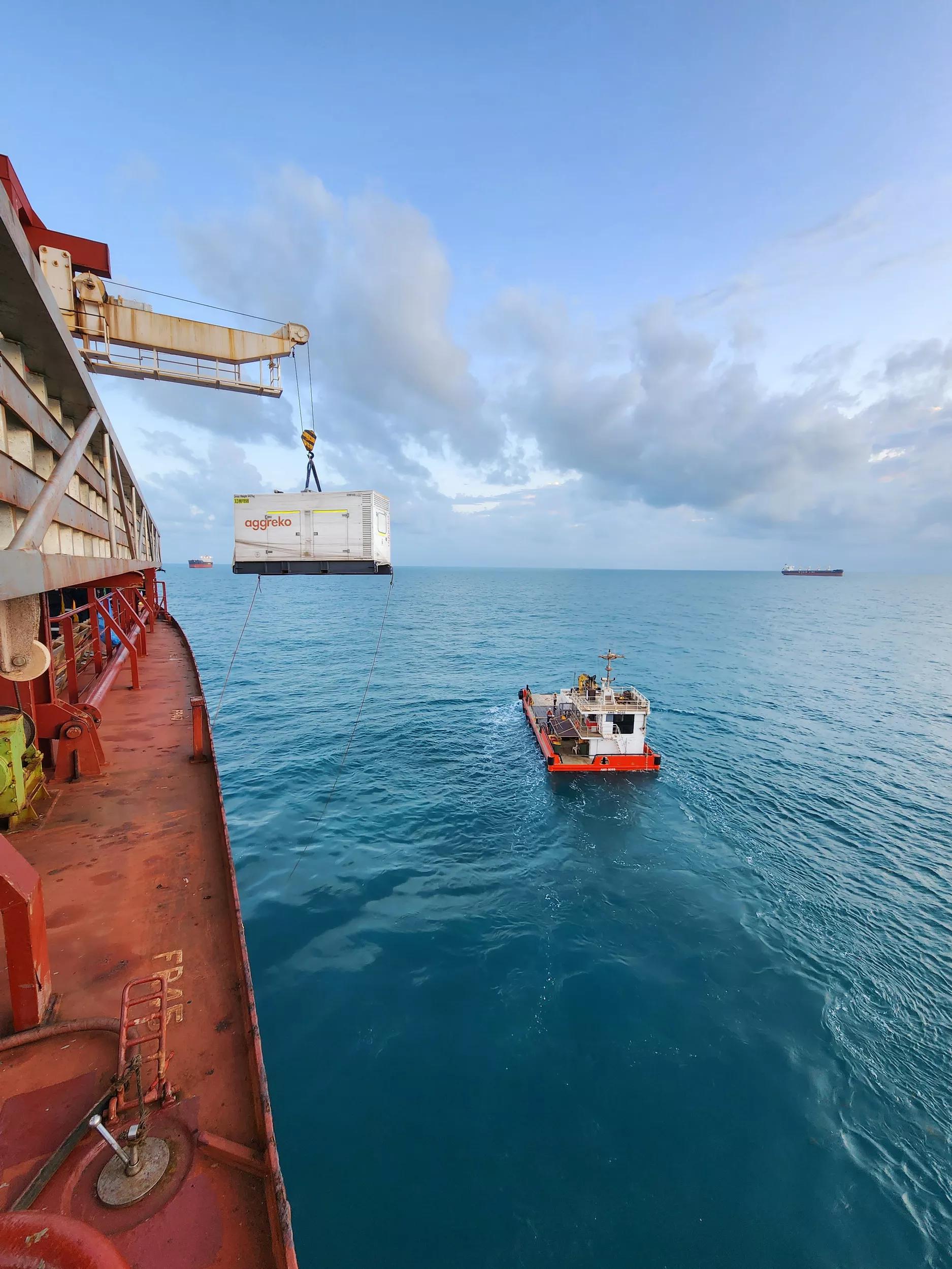 A white Aggreko generator is in front of red ships at a dock, with additional equipment and machinery nearby.