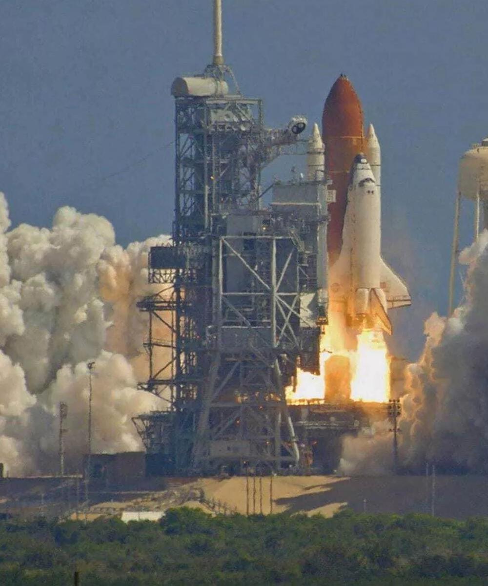 A space shuttle is captured mid-launch, rising vertically with massive plumes of fire and white smoke trailing below it. A tall water tower is visible on the right, and the scene is set against a bright, clear blue sky.