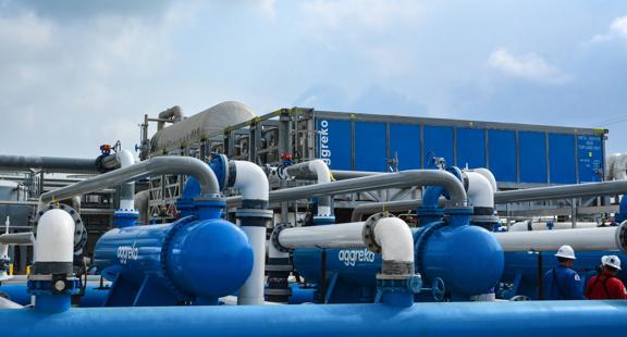 An extensive network of large blue and white industrial pumps and piping, including blue Aggreko branded horizontal vessels, set up at a facility with a large blue container unit visible in the background. Two workers in hard hats are visible near the piping.