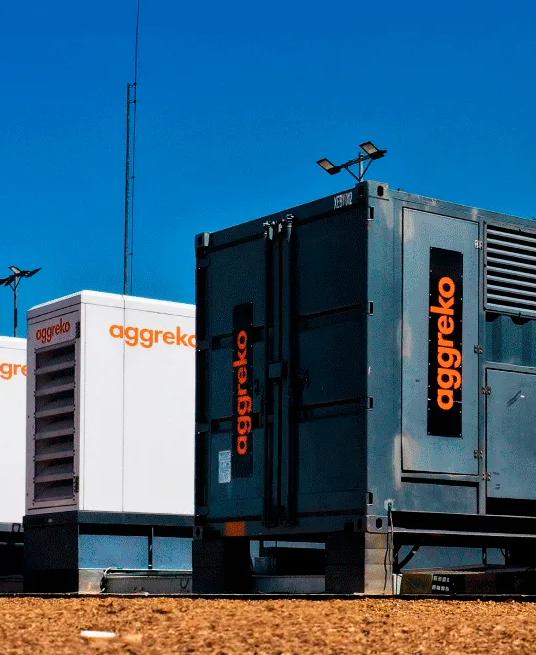 A row of Aggreko generator units on a dirt surface, with clear blue sky in the background.