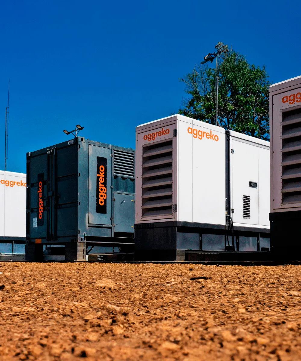 A row of Aggreko generator units on a gravel surface under a clear blue sky.