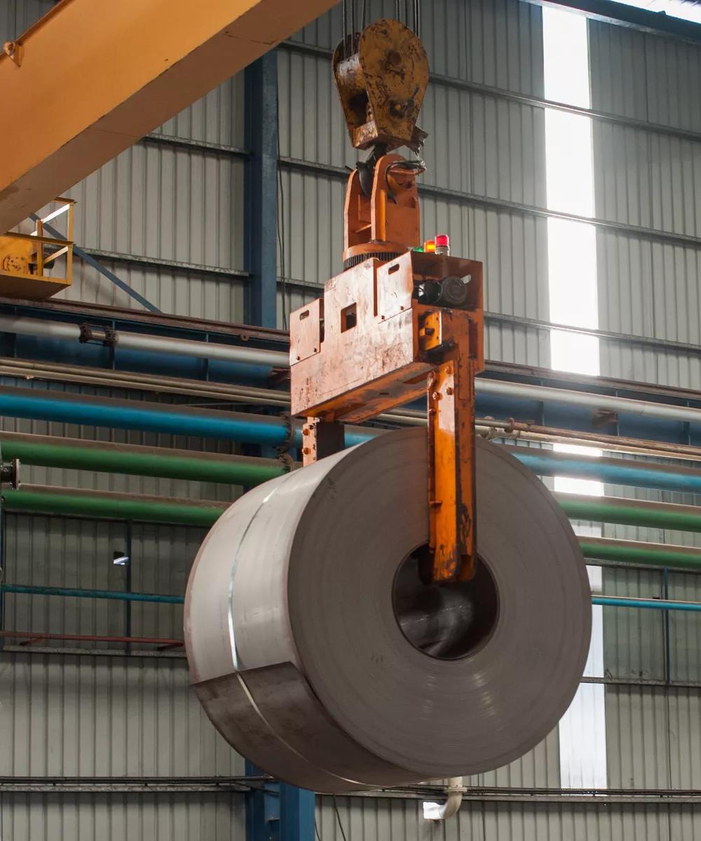 A dark, large roll of metal is suspended in a bright, modern factory setting. The coil is held by an orange overhead crane mechanism.
