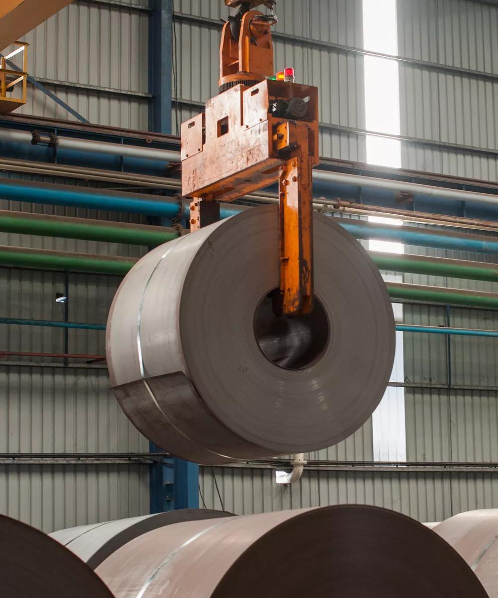 A dark, large roll of metal is suspended in a bright, modern factory setting. The coil is held by an orange overhead crane mechanism.