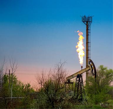 An oil rig with a flame burning at the top, surrounded by greenery and a cloudy sky.