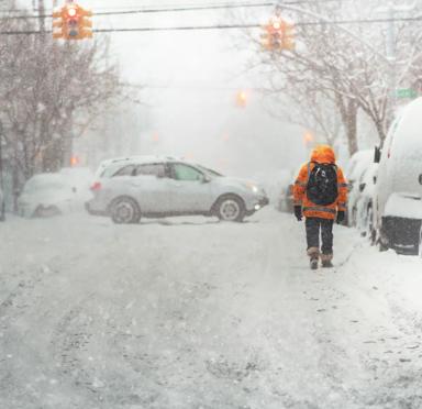 A person in a bright orange coat walks through heavy snow on a deserted street, with blurred cars and traffic lights in the background.