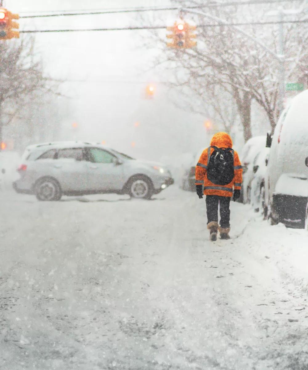 A person in a bright orange coat walks through heavy snow on a deserted street, with blurred cars and traffic lights in the background.