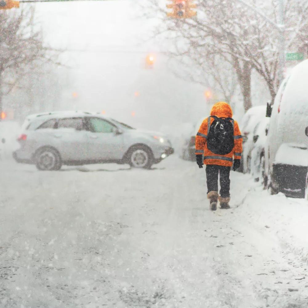 Person in an orange jacket walking down a heavily snowed street with cars parked beside and one turning around in the background.