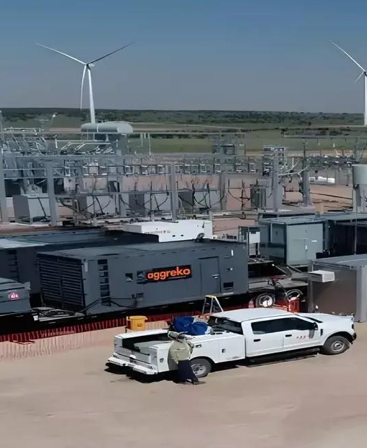 Aerial view of an industrial staging area next to an electrical substation. Several dark, containerized Aggreko industrial units are mounted on trailers on a gravel lot, with two white pickup trucks parked nearby in the dry, open landscape. Wind turbines visible in the distant background.