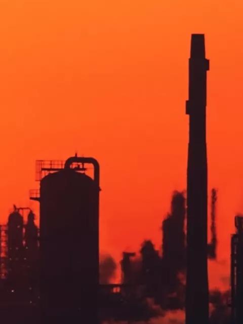 The silhouette of an industrial plant with smokestacks and pipes against a vibrant orange and red sunset.