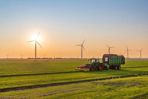 This is a photo of a tractor with a trailer harvesting on the agricultural field. In the background the sun is setting behind a row of wind turbines.
