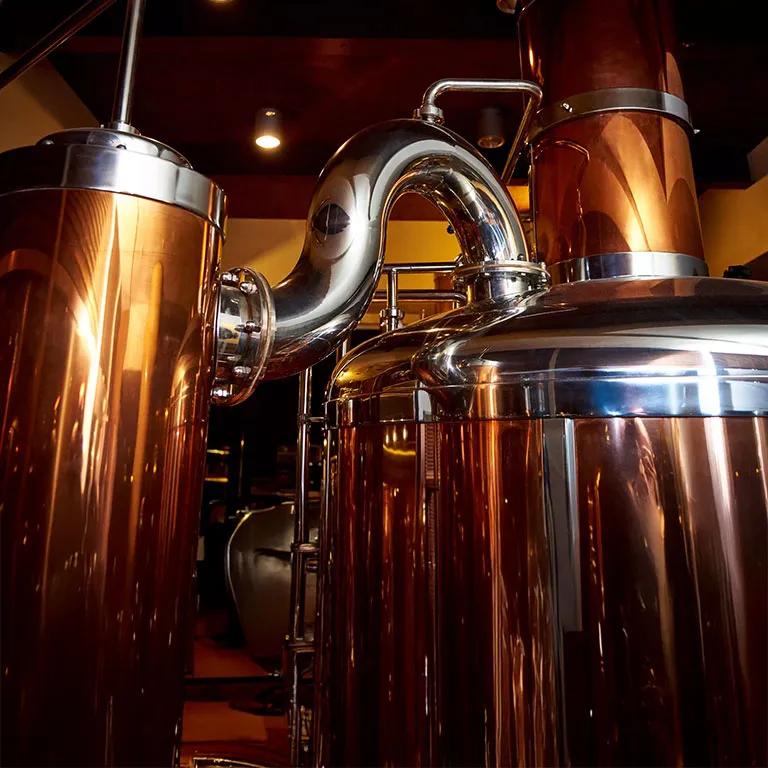 A close-up view of industrial brewing equipment, dominated by a large, curved, brightly polished stainless steel pipe connecting to a stainless steel tank. The background shows other large tanks with a copper or bronze sheen.