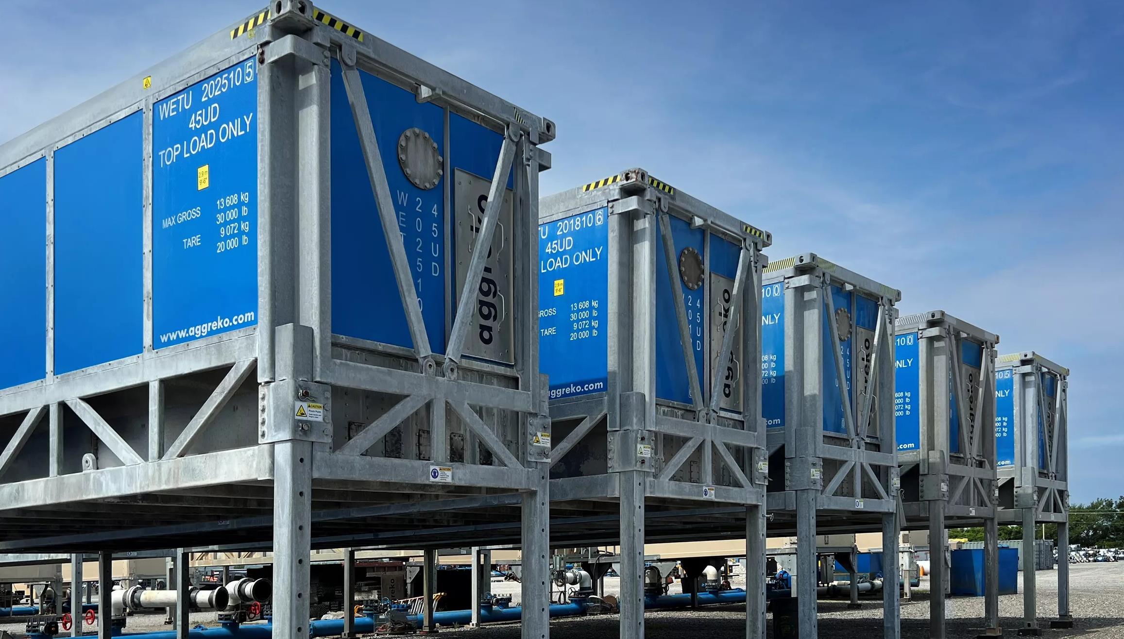 A line of big, blue industrial containers with gray metal frames, raised on legs under a blue sky.
