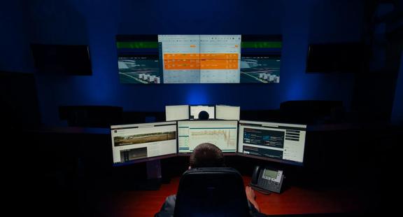 A dark control room with two workers seated at different desks with three computer monitors. Two screens in the background turned off. Two screens on the back, with a big central screen displaying performance metrics.