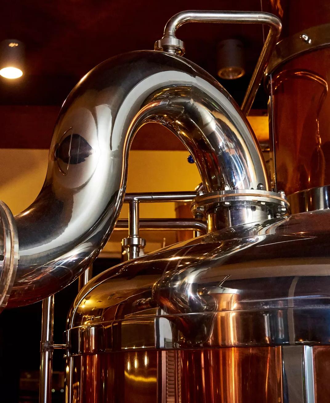 A close-up view of industrial brewing equipment, dominated by a large, curved, brightly polished stainless steel pipe connecting to a stainless steel tank. The background shows other large tanks with a copper or bronze sheen.