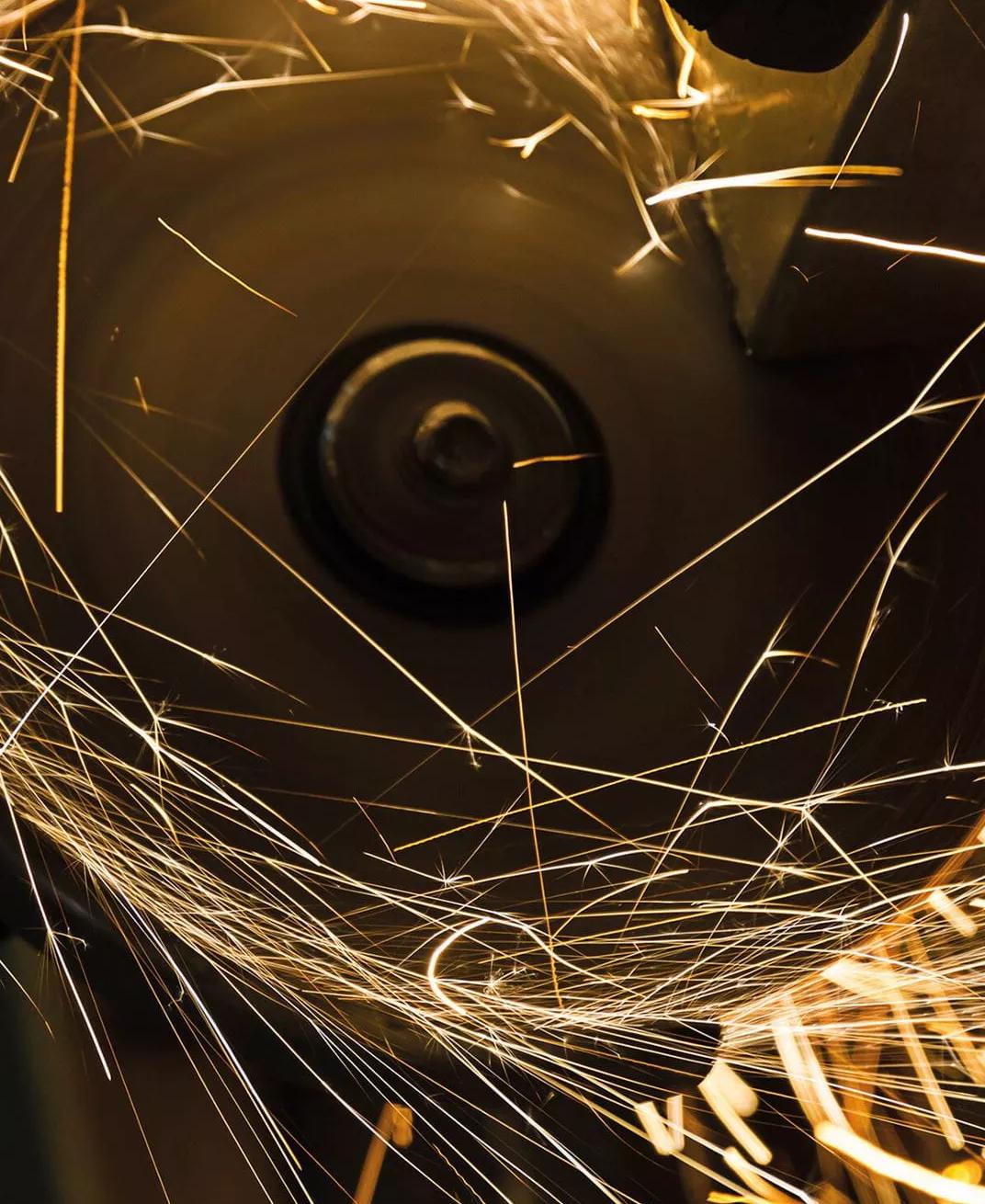 A dark, close-up shot capturing a cascade of bright orange and yellow sparks or embers flying off a piece of metal.
