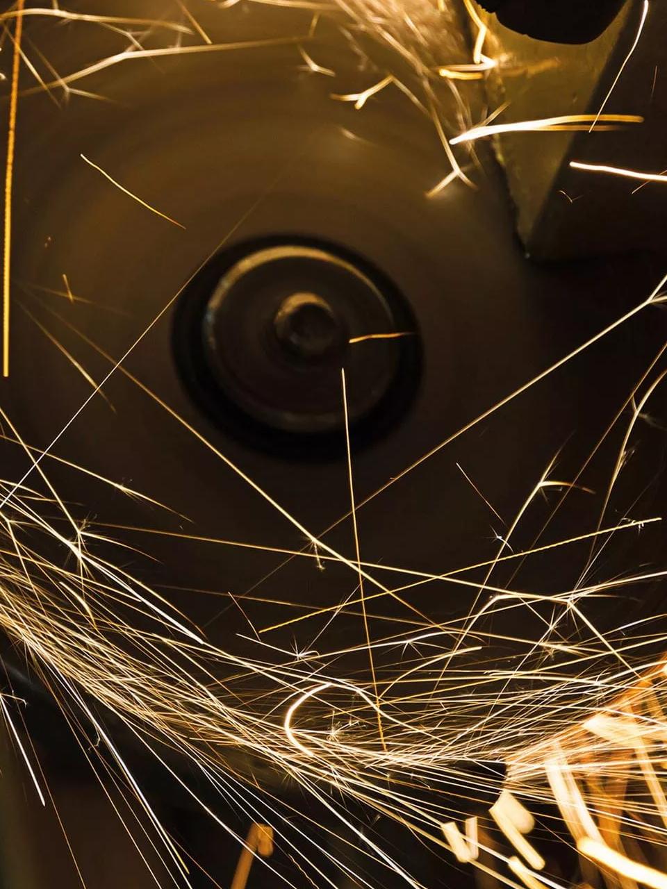 A dark, close-up shot capturing a cascade of bright orange and yellow sparks or embers flying off a piece of metal.