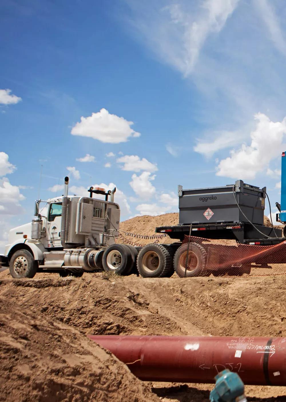 A blue, containerized Aggreko generator is mounted on a dual-axle trailer in a barren, dusty outdoor location under a bright blue sky.